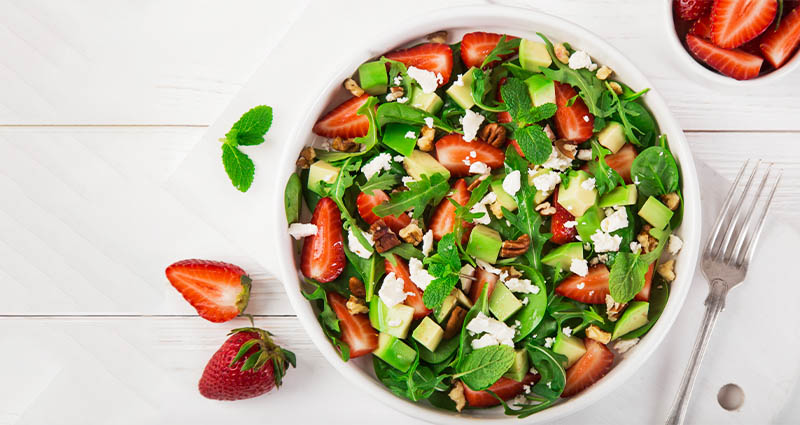  A vibrant salad in a large white bowl, featuring mixed greens, diced fruits, and nuts, with fresh strawberries placed beside it on a light wooden surface.