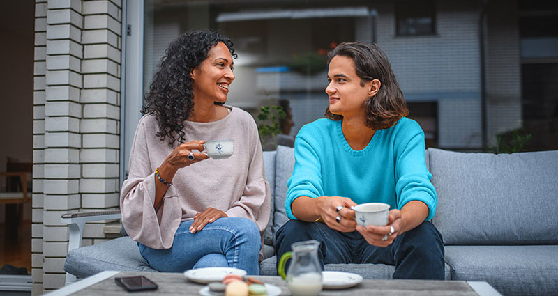 Two women are sitting on a couch outdoors, smiling and enjoying coffee together, engaged in a friendly conversation.