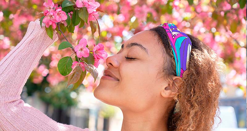 A young woman with curly hair and a colorful headband joyfully smelling pink flowers in bloom, with a soft focus background of a flower-laden tree.