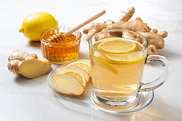 A glass of ginger tea with lemon slices. Nearby are fresh ginger, lemon, a honey dipper, and a bowl of honey on a white surface.