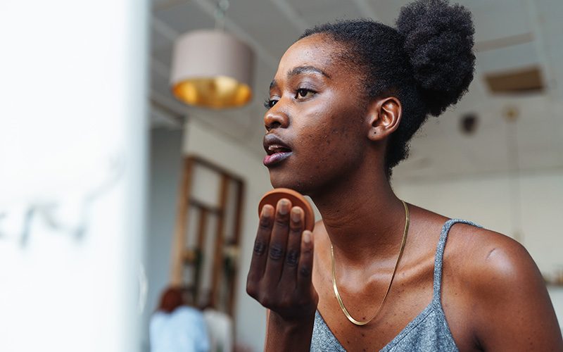 Young woman applying makeup with a sponge while looking in a mirror.