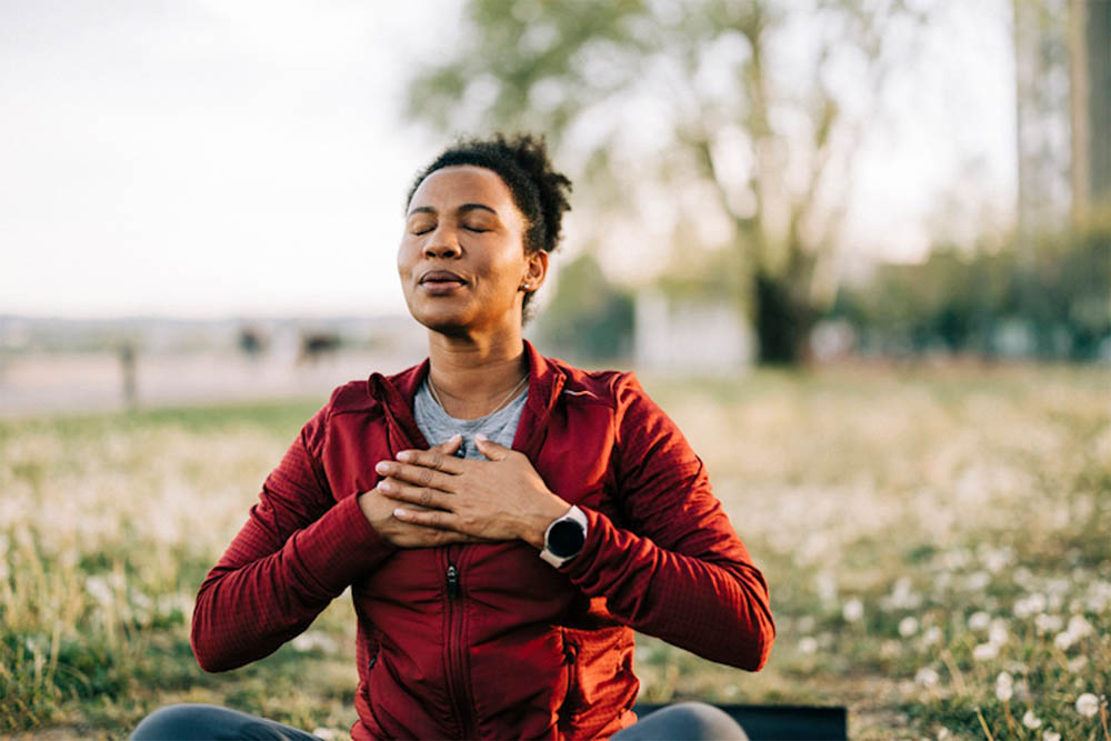 Woman sitting cross-legged in a field, practicing deep breathing with her hands on her chest.