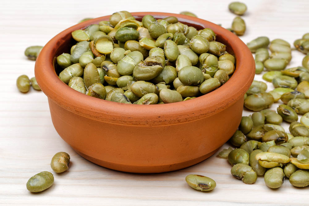A clay bowl filled with roasted green soybeans, some spilled on a light wooden surface.