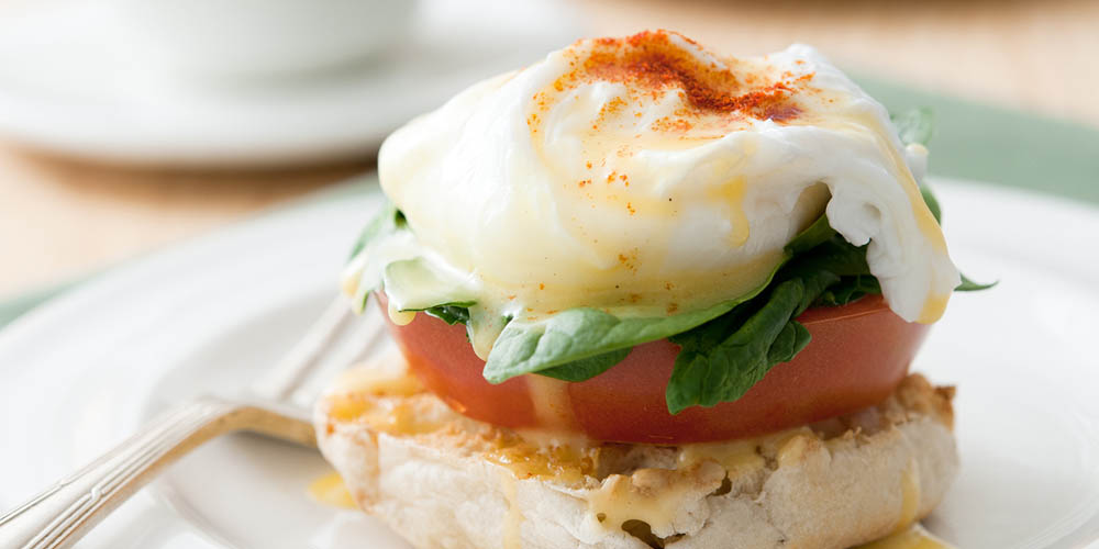 A close-up of a toasted bread slice topped with spinach, a slice of tomato, and a poached egg, served on a plate with a fork beside it.