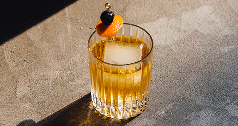 Nonalcoholic old fashioned cocktail with a large ice cube and cherry/orange garnish in a rocks glass, sitting on a marble countertop.