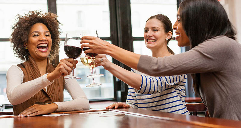 Three women doing a cheers with wine glasses while sitting at a wooden table or bar, looking celebratory.