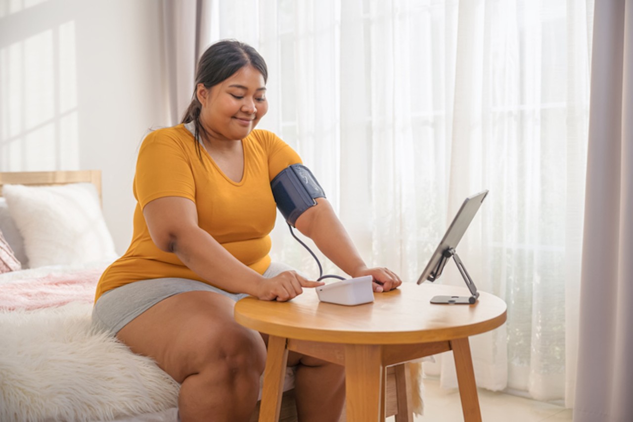 A person sits on a bed measuring their blood pressure with a device on a wooden table, wearing a yellow shirt and gray shorts, in a bright room.