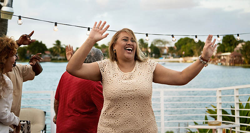 A joyful woman dancing outdoors at a social gathering near the water, surrounded by friends and festive string lights.