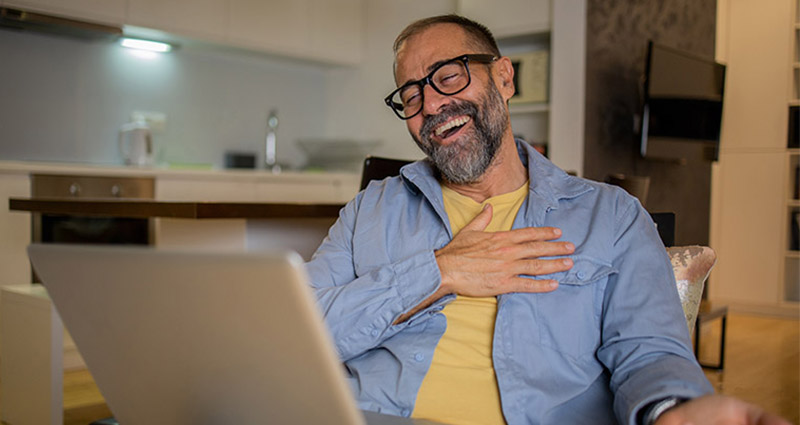 A middle-aged man with glasses and a beard smiling warmly while engaging in a virtual therapy session on his laptop at home.