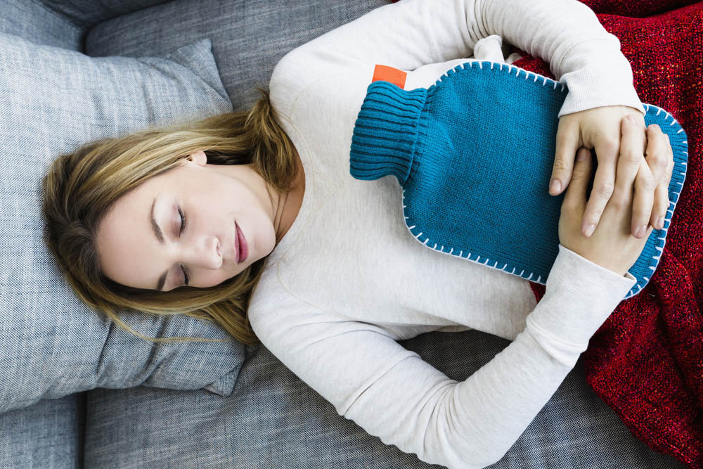 A person lies on a couch with eyes closed, holding a blue hot water bottle on their midsection, covered by a red blanket.