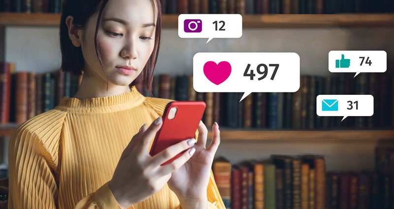 A woman is looking at her smartphone with various social media notifications displayed, including likes and comments, against a background of bookshelves.