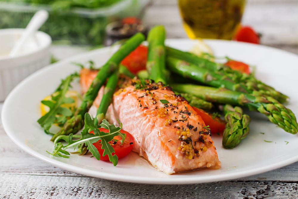 A plate of grilled salmon fillet served with asparagus and cherry tomatoes, garnished with fresh greens, and accompanied by a small bowl of sauce in the background.