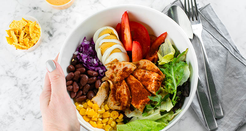  A close-up of a colorful salad bowl being held by a hand. The bowl contains various ingredients, including grilled chicken, black beans, corn, sliced eggs, lettuce, and red cabbage, arranged beautifully