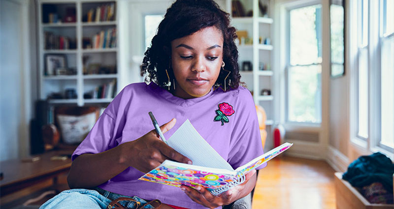 A young woman in a purple shirt is sitting at a table, writing in a colorful notebook, with a cozy room and bookshelves visible in the background.