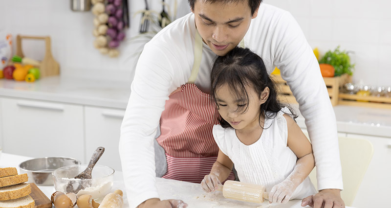 A parent and child are engaged in baking in a kitchen, with the parent guiding the child as they roll out dough on a countertop, surrounded by baking ingredients and utensils.