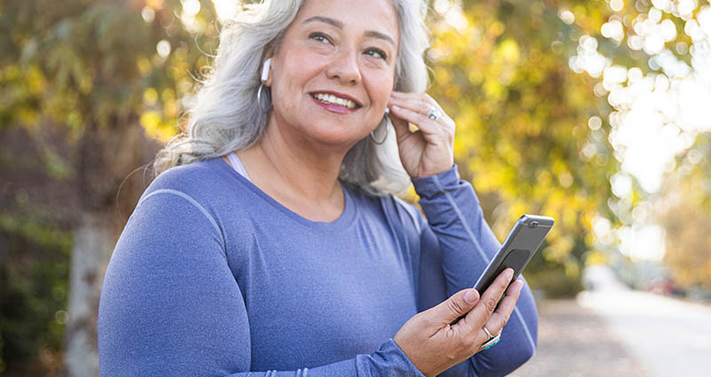 A smiling woman with long gray hair is outdoors, wearing wireless earbuds and holding a smartphone, dressed in a long-sleeved athletic shirt, with trees in the background.