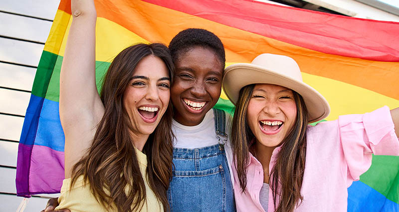 Three smiling women pose together, holding a colorful rainbow pride flag, celebrating with joy, in a bright and cheerful atmosphere.