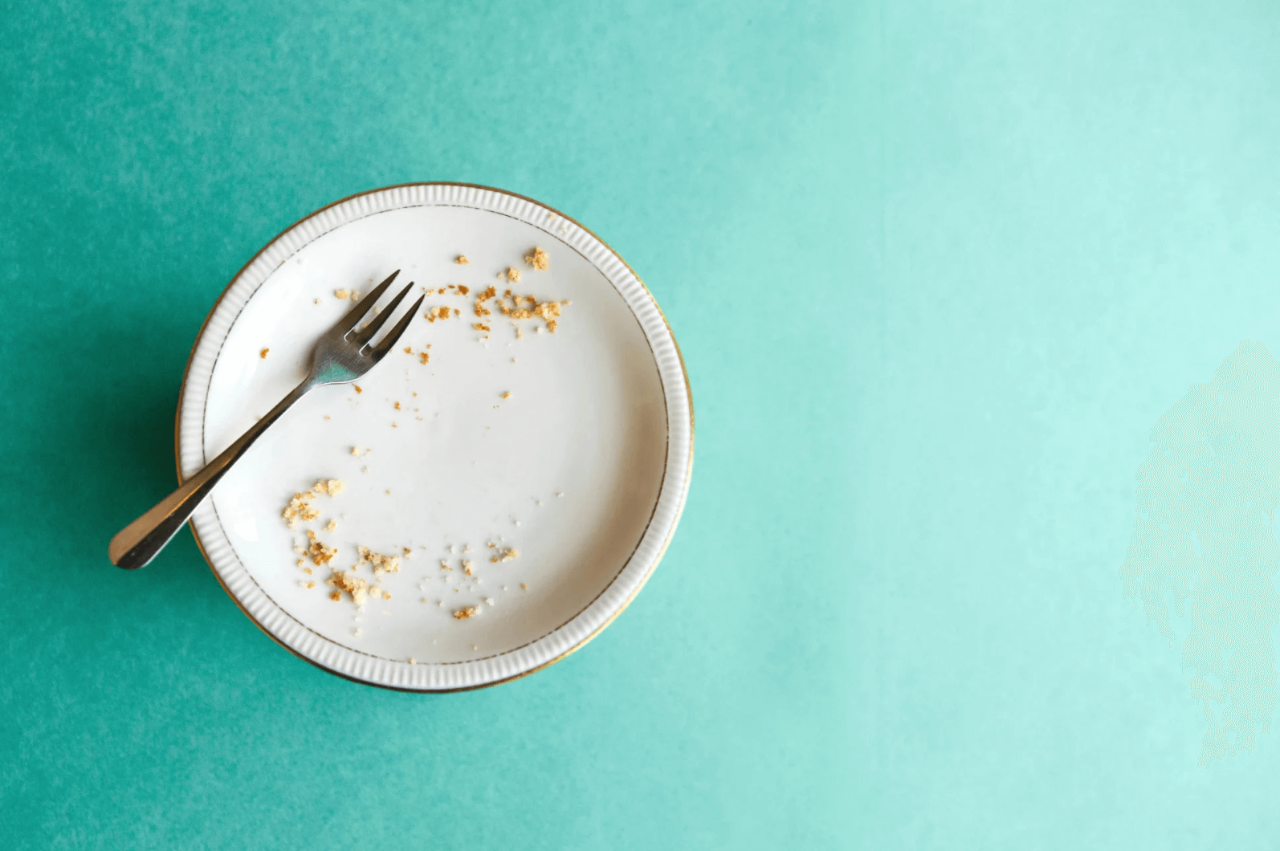 Empty white plate with a fork, crumbs scattered on the surface. Set against a solid turquoise background.