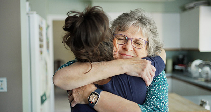 An elderly woman with glasses embraces a younger woman in a warm hug inside a kitchen.