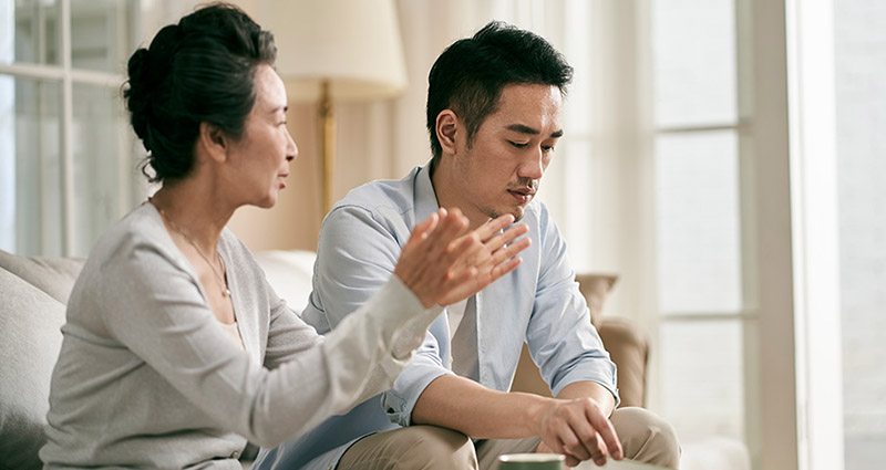 A concerned older woman gestures while speaking to a younger man who looks down, appearing deep in thought. They are sitting in a bright living room.