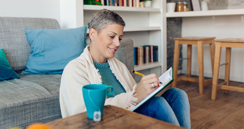 A smiling woman sitting on the floor, wrapped in a blanket, writing in a journal with a coffee cup beside her in a cozy living room setting.
