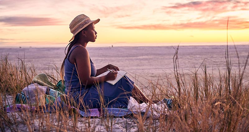 A woman in a blue dress and straw hat sits on a blanket in the dunes, journaling while watching the sunset over the beach.