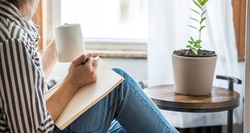 A person sitting by a window, holding a coffee mug and writing in a journal, with a potted plant on a wooden side table nearby.