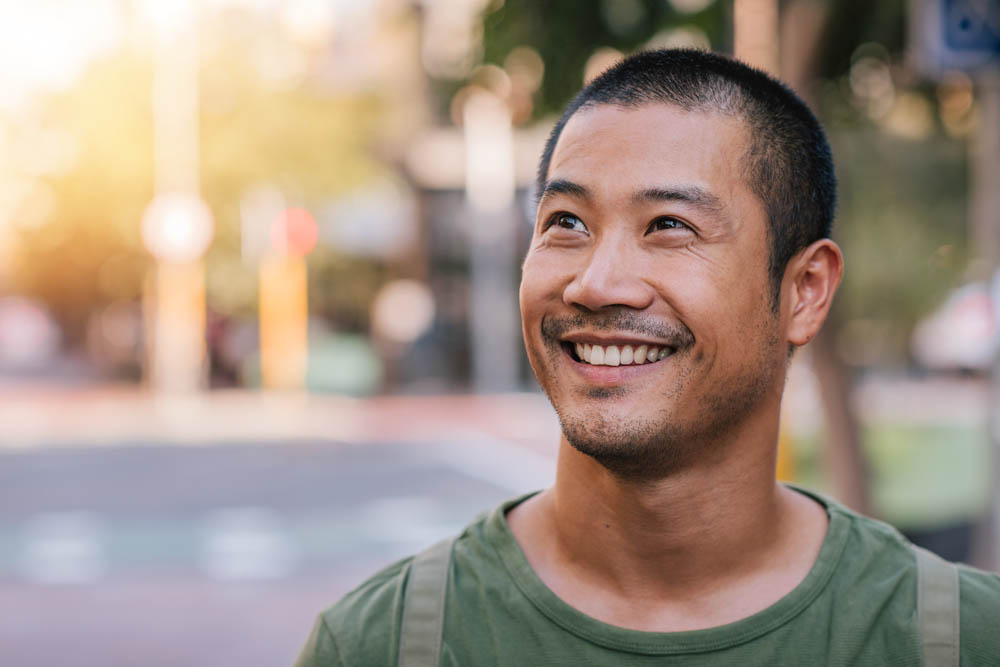 Casually dressed handsome young Asian man looking up and smiling while standing alone outside on a city street on a sunny day