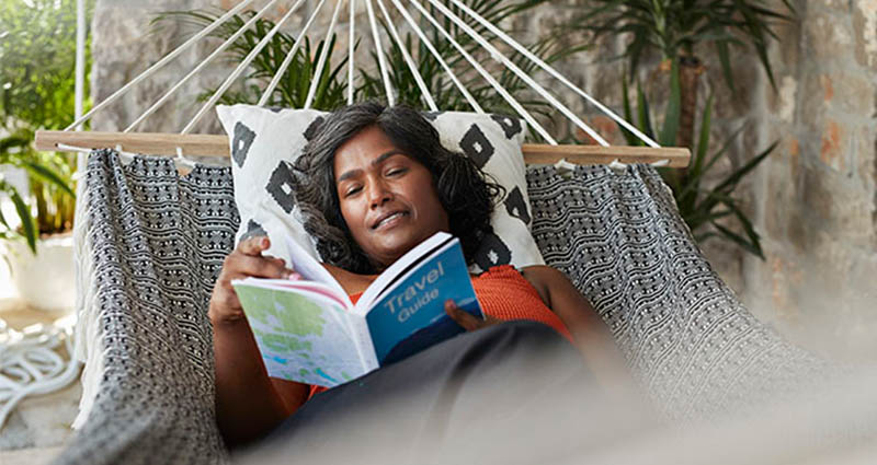 A woman relaxing in a hammock, reading a travel guide, with cozy pillows and lush greenery around her.