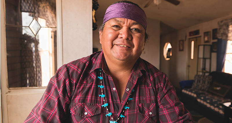 A smiling man wearing a purple headband, a plaid shirt, and a beaded necklace, sitting in a warmly lit home.