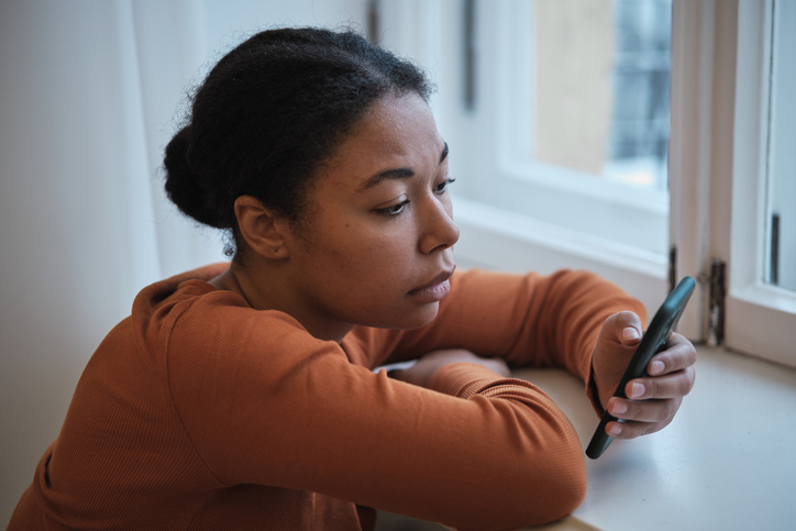 Woman sitting by a window looking at her phone with a thoughtful expression