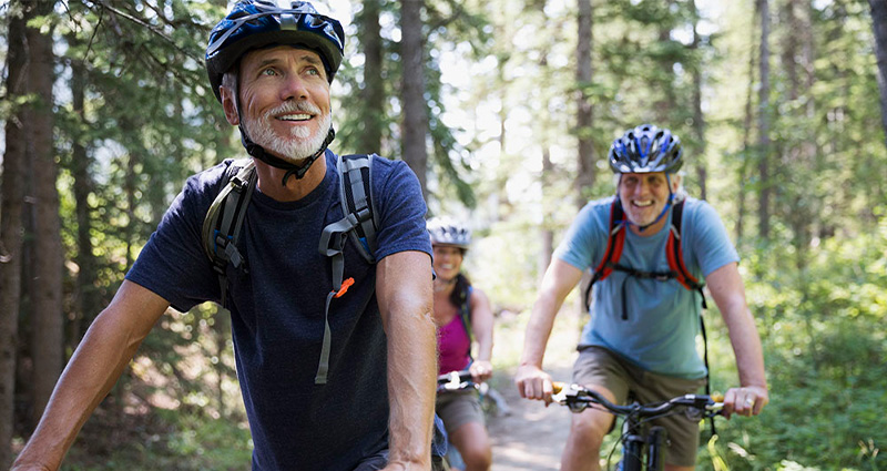 A group of three older adults riding bicycles through a forest trail, smiling and enjoying the outdoors.