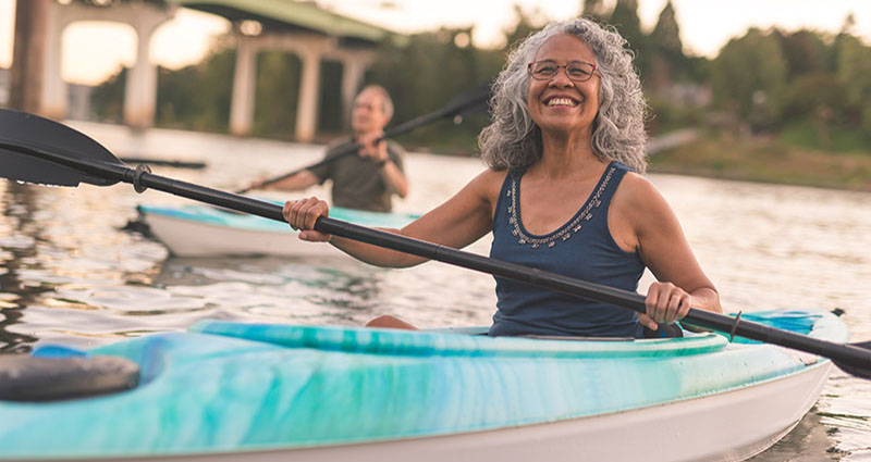A smiling older woman with curly gray hair kayaking on a river, with another person paddling in the background. The setting sun casts a warm glow on the water.