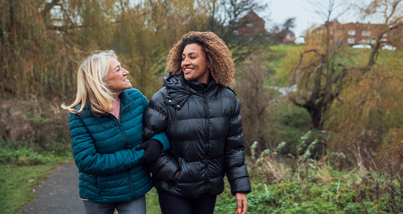 Two women walking outdoors on a paved path, smiling at each other. One woman wears a blue winter coat, and the other wears a black puffer jacket