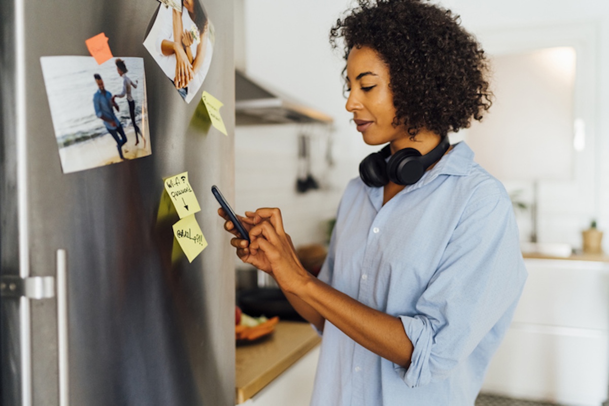 Woman standing in a kitchen, using her phone with headphones around her neck and sticky notes on the refrigerator.