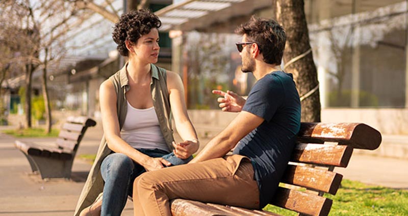 A man and a woman sitting on a wooden bench in a park, engaged in a serious conversation.