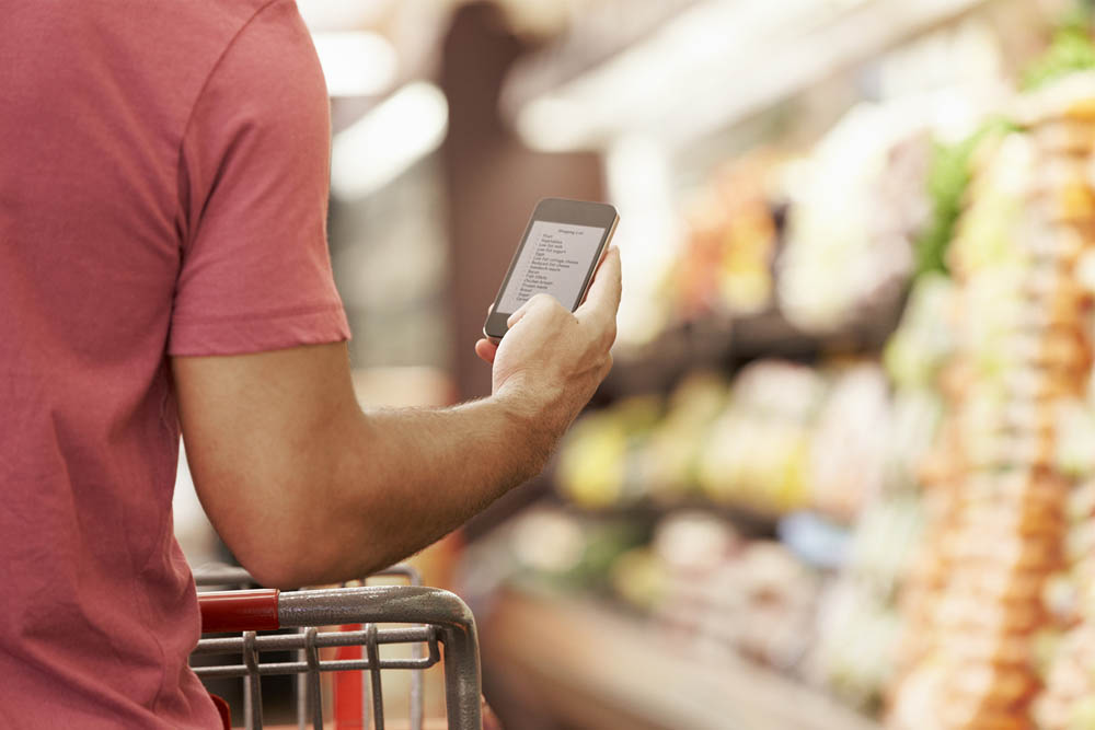 A person is holding a smartphone while pushing a shopping cart in a grocery store, with shelves of colorful produce in the background.