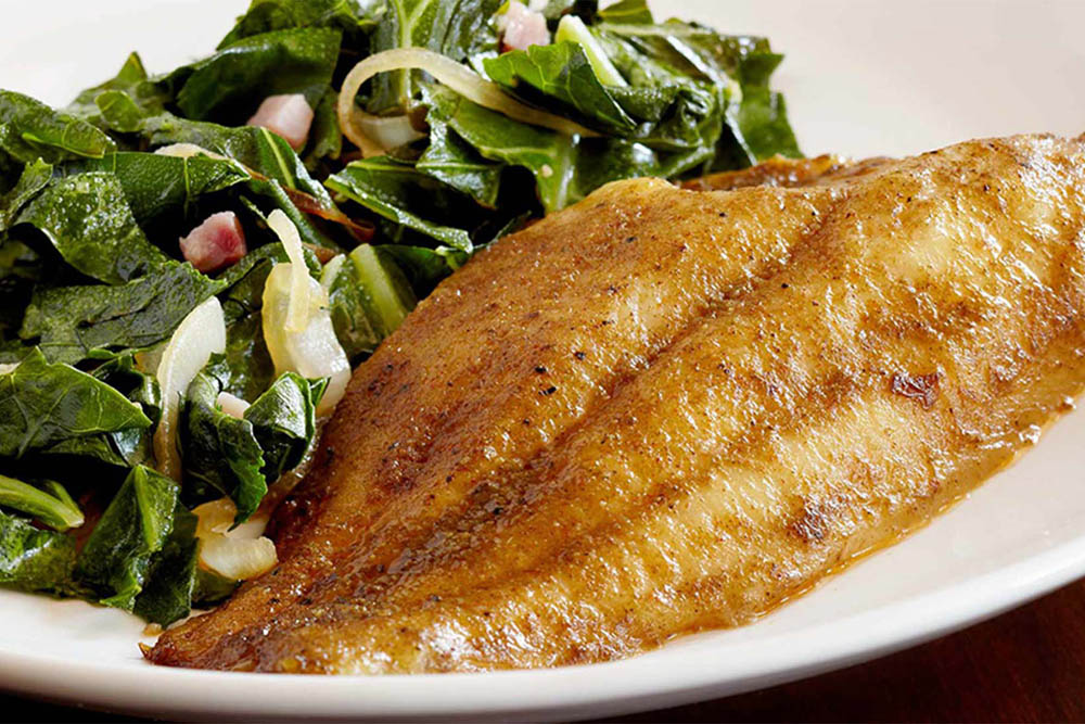  A plate featuring a fillet of fried fish alongside a serving of sautéed greens, with hints of onion visible in the greens, all presented on a white dish.