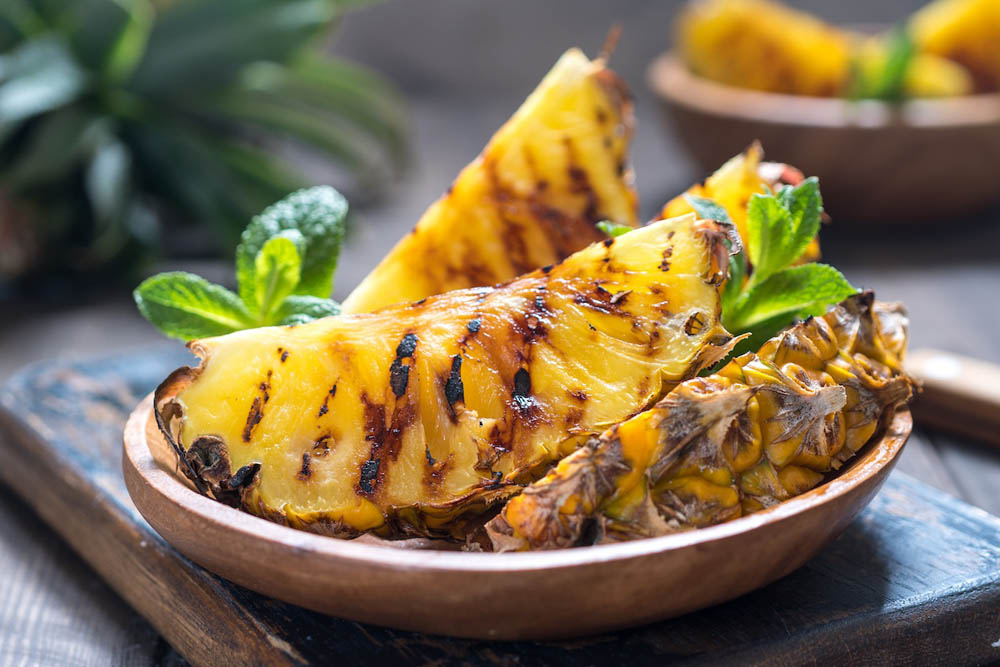 A wooden bowl filled with grilled pineapple slices garnished with mint leaves, set against a rustic background with hints of fresh pineapple in the backdrop.