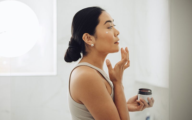 Woman applying facial moisturizer while looking in the mirror.