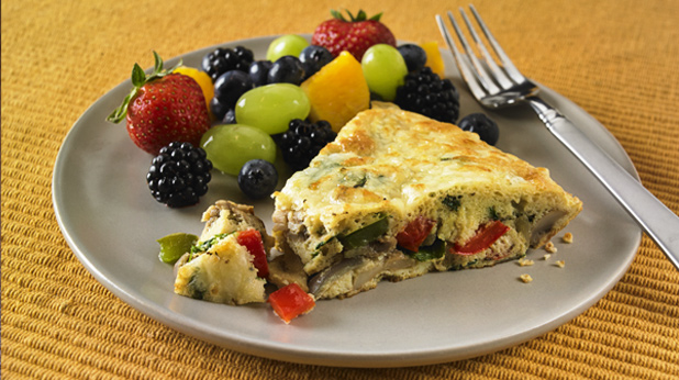A plate featuring a slice of vegetable frittata alongside a colorful assortment of fresh berries and grapes, with a fork resting on the side, set against a textured yellow background.