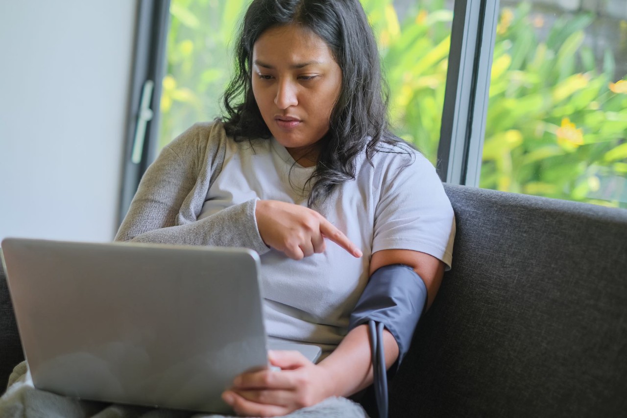 Close-up shot of sick Asian woman showing blood pressure gauge on her arm to her online doctor. She's on video conference via laptop.