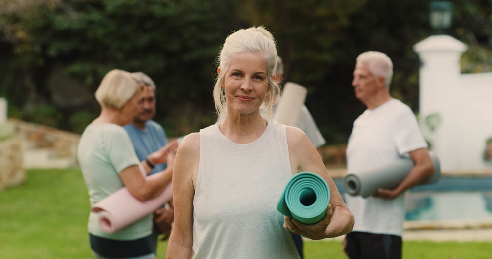 A woman holding a yoga mat standing in the foreground. Two other people with yoga mats converse in the background, outdoors.