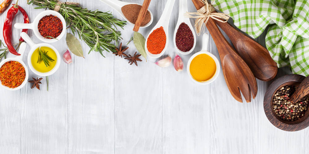Various spices, herbs, and wooden utensils are arranged on a white surface with a green checkered cloth to the side.