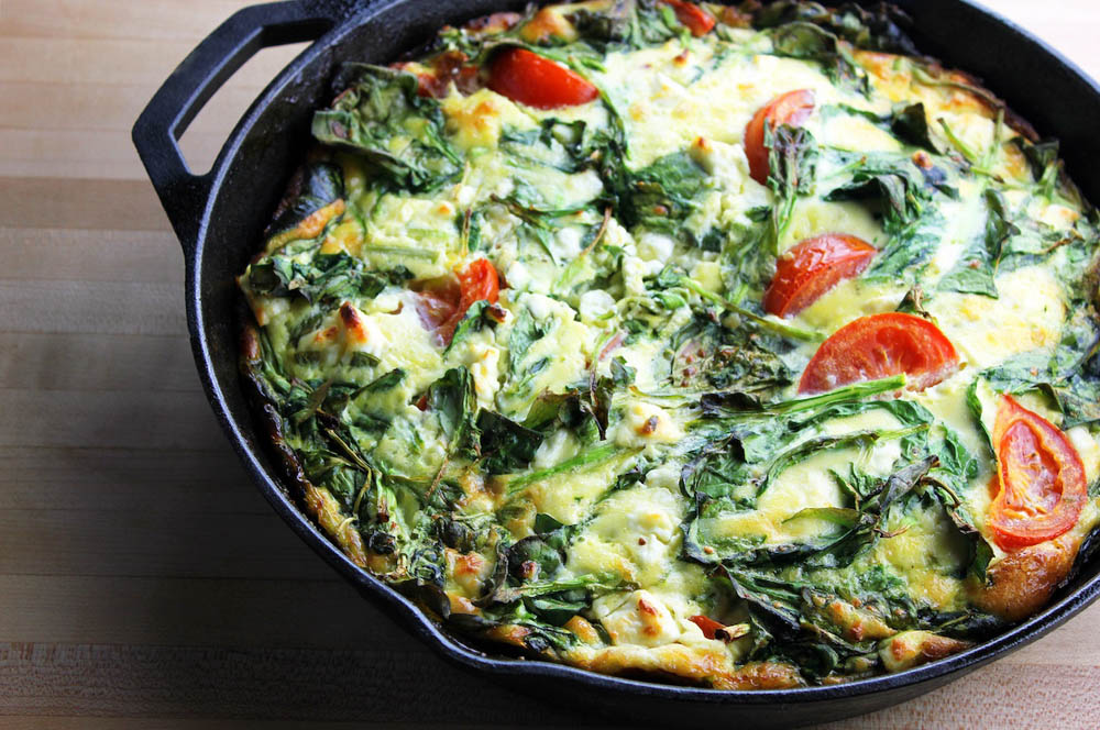 A cast iron skillet filled with a baked frittata, featuring a mix of greens and sliced tomatoes, resting on a wooden surface.