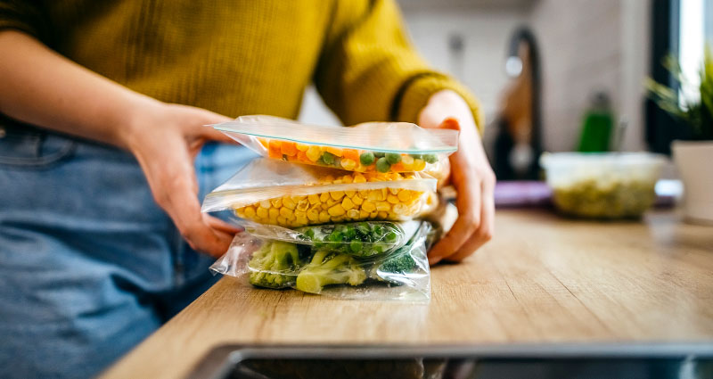 Person organizing frozen vegetables in airtight bags on a wooden countertop.