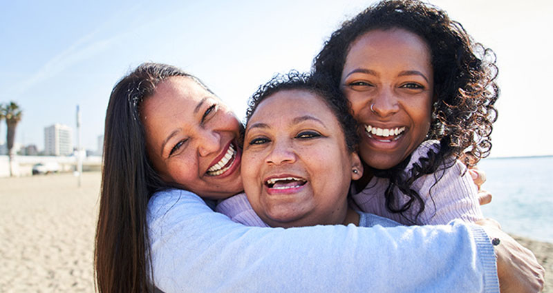 Three women smiling and hugging each other on a sunny beach.