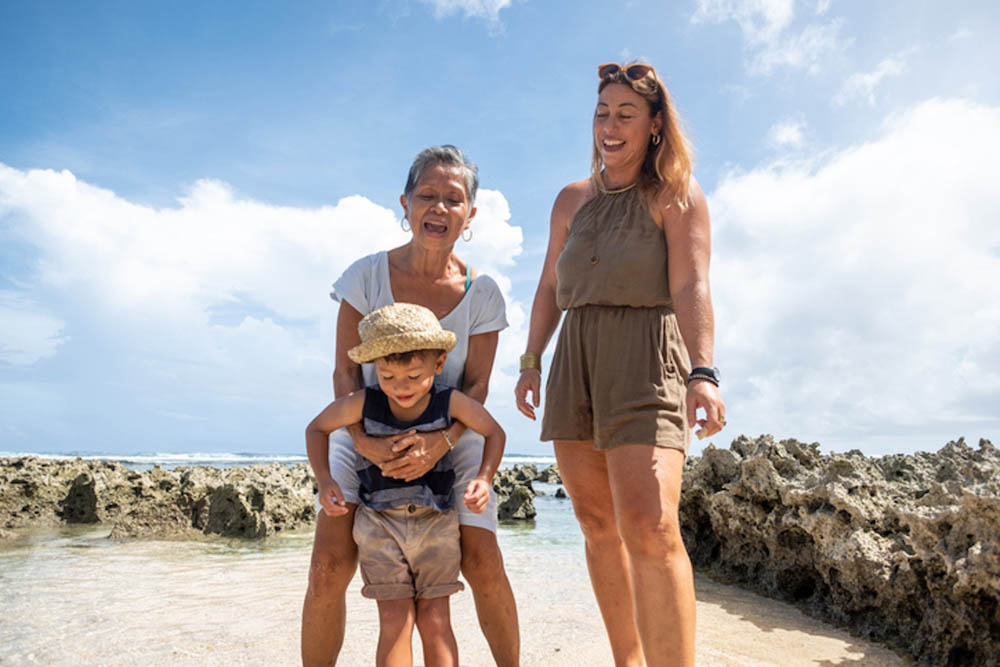 Three generations of a family enjoying a day at the beach, smiling under a bright sky.