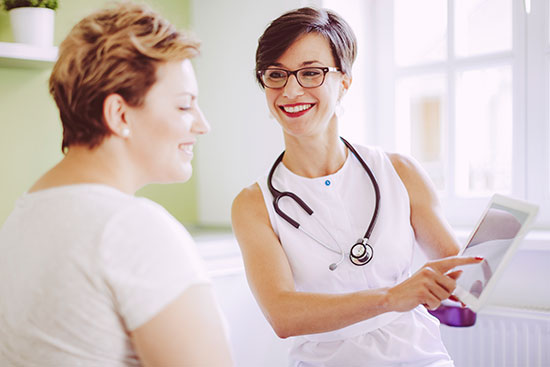 A smiling doctor with a stethoscope showing a digital tablet to a seated patient in a well-lit room.