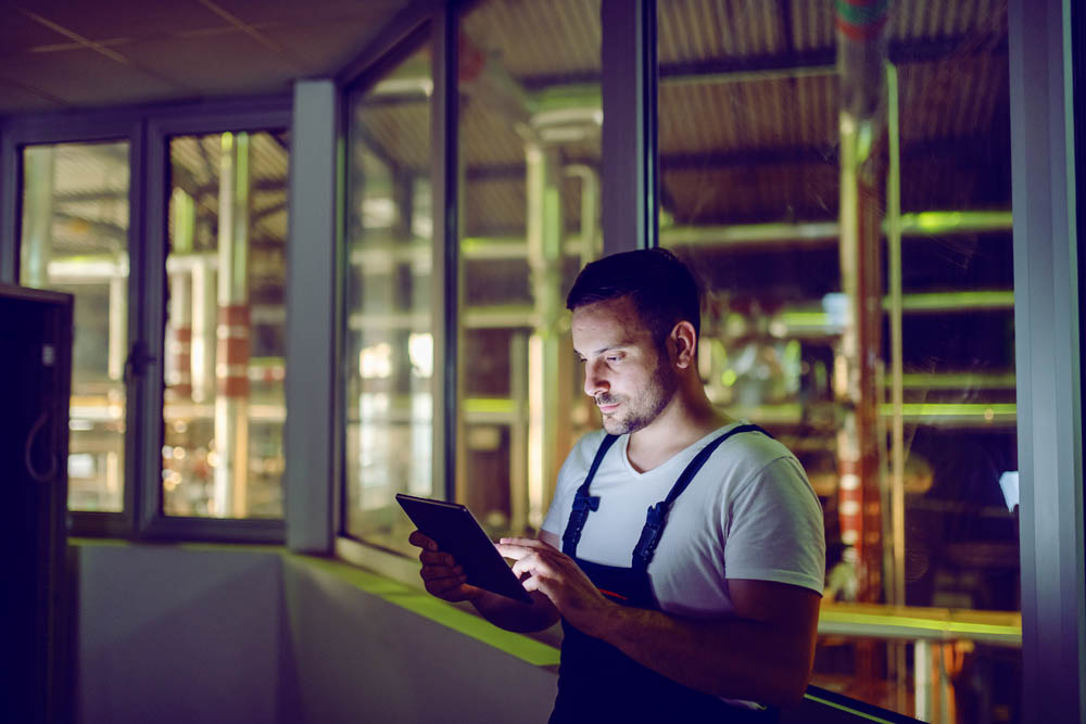 Hardworking plant worker in coveralls standing and using tablet at night. Night shift.
